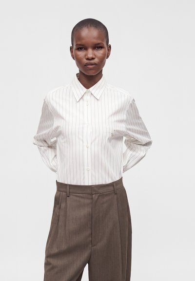 Personne portant une chemise blanche à fines rayures boutonnée et un pantalon plissé marron taille haute, debout les mains dans le dos.