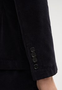 Manche de blazer en velours côtelé bleu marine foncé avec nervures verticales, présentant quatre boutons noirs et un bord de manche lisse.