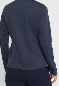 Námorně modrá sportovní bunda vyrobená z měkké, texturované tkaniny s dlouhými rukávy a přiléhavým střihem; má kulatý výstřih a minimální švy.