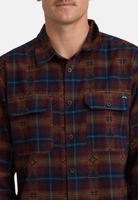 Langsleeve geruite overhemd in roest, marineblauw en groen. Bevat twee borstzakken, knoopsluiting en een textuurstof met geometrische patronen.