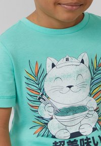 Turquoise katoenen t-shirt met een cartoonkat die een kom noedels met een eetstokje vasthoudt, omringd door kleurrijke bladerenpatronen.