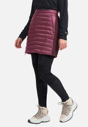 Lichtgewicht, geïsoleerde, bordeauxrode rok met een gewatteerd ontwerp en een zijrits; gecombineerd met zwarte leggings en beige wandelschoenen.