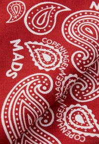 Rød stoff med hvite paisleymønstre og tekst som sier "MADS COPENHAGEN." Materialet ser mykt ut med en glatt tekstur.