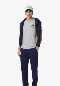 Pantalones de chándal azul marino y sudadera sin mangas sobre una camiseta gris con un logo. El modelo lleva una gorra gris claro. Tejido suave, estilo atlético casual.