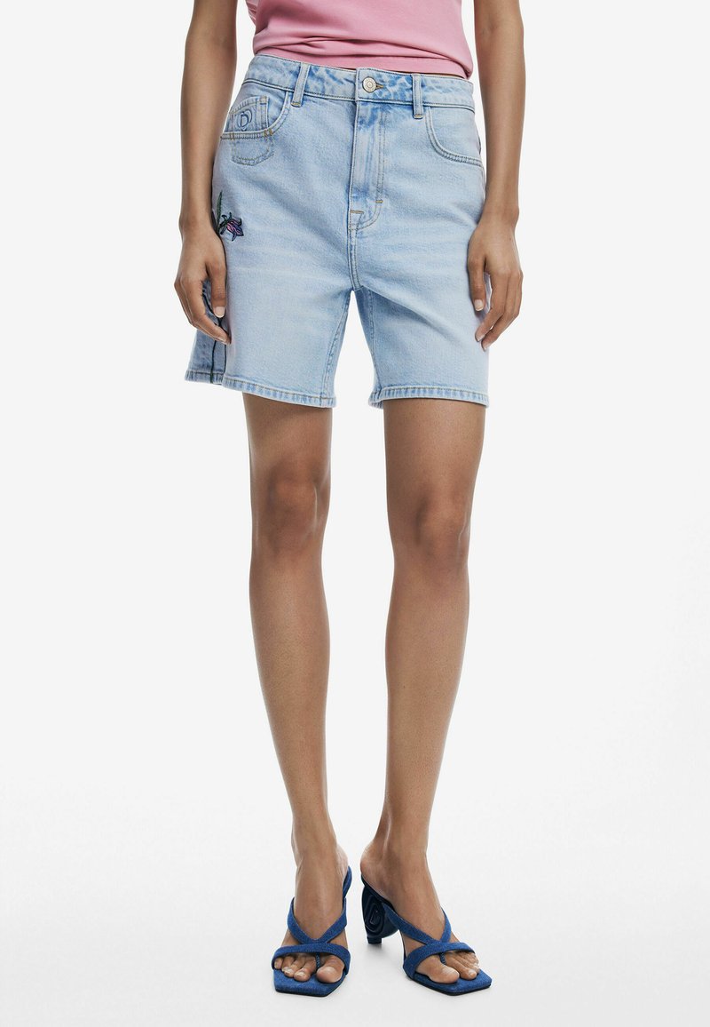 Shorts de mezclilla azul claro con un corte relajado, que presentan un detalle de bordado floral en la pierna izquierda y un cierre de botón en la cintura.