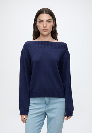 Femme aux longs cheveux foncés, portant un pull bleu marine à épaules découvertes et un jean bleu clair, debout devant un fond blanc uni.