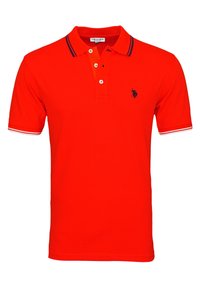 Punainen polo-paita lyhyillä hihoilla, kolmellä napilla varustettu nappilista ja pieni tummansininen emblem vasemmassa rinnassa. Sissä on tummansiniraidallinen ja valkoinen kaulus.