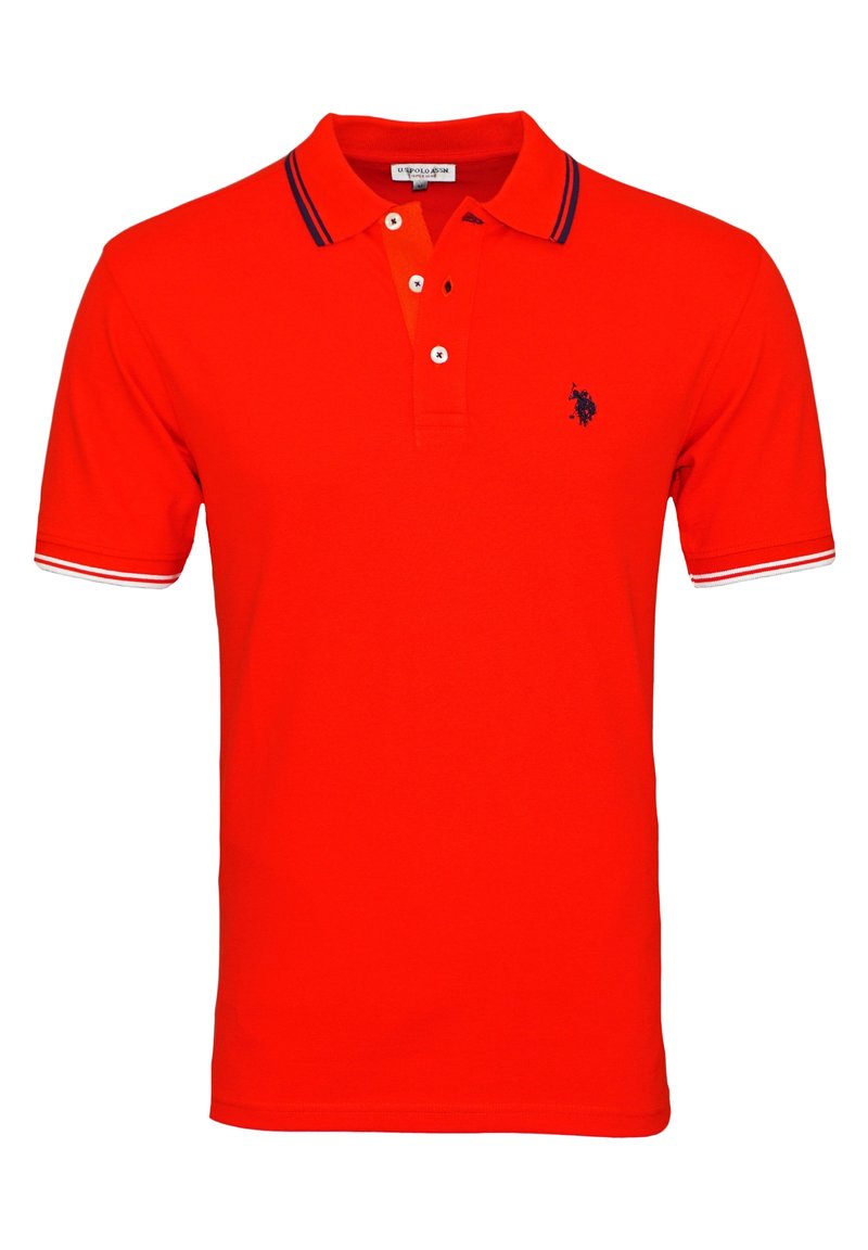 Punainen polo-paita lyhyillä hihoilla, kolmellä napilla varustettu nappilista ja pieni tummansininen emblem vasemmassa rinnassa. Sissä on tummansiniraidallinen ja valkoinen kaulus.