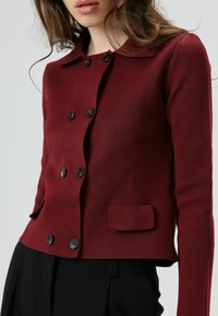 Veste en maille rouge avec col arrondi, boutons noirs croisés et petite poche avant. Texture lisse et coupe ajustée.