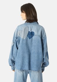 Lys blå denimjakke med oversized passform. Den har applikert hjertepatcher i forskjellige nyanser, slitt hem og knappelukking.
