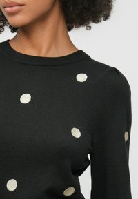 Suéter de punto negro con puntos metálicos dorados, cuello redondo y sutiles mangas abullonadas. Textura suave con un estilo ajustado.