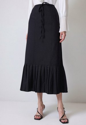 Gonna nera maxi con vita regolabile, caratterizzata da un orlo plissettato e tessuto uniforme. Abbinata a una maglietta bianca a maniche lunghe e sandali neri.