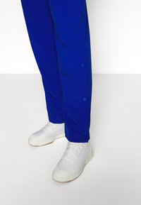 Blauwe broek met drukknoopjes aan de zoom, gecombineerd met witte sportschoenen met een gestructureerde zool en vetersluiting.