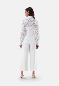 White lace blouse with floral pattern, long sleeves, and collar, paired with white wide-leg pants. Model stands showing back view.
