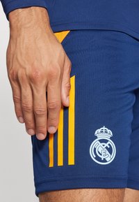 Siniset urheilushortsit keltaisilla pystysuorilla raidoilla, joissa on valkoinen Real Madrid -logo alhaalla oikealla puolella. Pehmeä, joustava kangas.