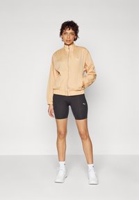Veste coupe-vent pêche clair avec un col montant et une fermeture éclair, associée à un short de cyclisme noir, tous deux arborant de subtils accents de logo.