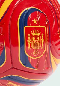 Krupni plan crveno-žutog fudbalskog lopte s prikazom nacionalnog grba Španije koji sadrži krunu, stubove i emblem lava.