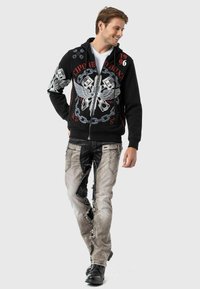 Schwarze Zip-Hoodie mit grafischen Drucken und Kettendesign, kombiniert mit ausgewaschenen grauen Jeans mit Patches und schwarzen Akzenten.