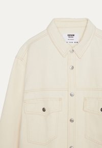 Krämfärgad denimjacka med krage, silverfärgade tryckknappar och två framfickor. Synlig sömnad i en kontrasterande färg.