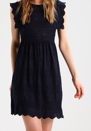 Kobieta w granatowej sukience z krótkim rękawem, ozdobionej koronką z ażurowymi wzorami i falbanką na dole, stojąca na jednolitym tle.