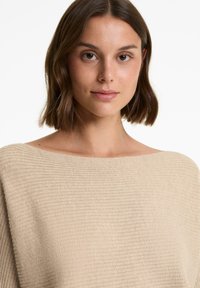 Beige bordázott kötött felső széles hajónyakkivágással, sima textúrával és laza fazonnal, amelyet rövid barna hajú modell visel egy semleges háttér előtt.