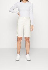 Pantalones cortos beige hasta la rodilla en un diseño sólido, hechos de una tela ligera, combinados con una camiseta de manga larga color gris claro y zapatillas blancas.