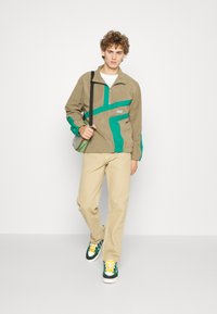Chaqueta beige con media cremallera y acentos diagonales verdes, combinada con pantalones caqui claros y zapatillas de colores. Llevando una pequeña bolsa cruzada.