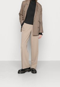 Blazer oversize a scacchi in marrone e beige, abbinato a un dolcevita nero a coste e pantaloni beige sartoriali. Scarpe con plateau nere.