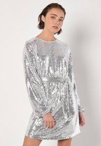 Robe en sequins argentés avec des manches longues et un col rond, équipée d'une ceinture à la taille. Texture lisse avec une surface réfléchissante.