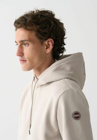 Béžová mikina s kapucí a šňůrkami, jemná textura a kulatý logový nášivka na rukávu. Jednoduchý design s velkou kapucí pro ochranu.