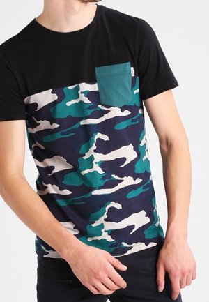 Mand iført sort T-shirt med korte ærmer, der har et camouflage-mønster i turkis, marineblå og hvid samt en ensfarvet turkis brystlomme.