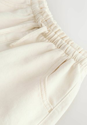 Gros plan sur un pantalon couleur crème avec une taille élastique, un cordon de serrage et des coutures visibles sur la poche avant, sur un fond clair.