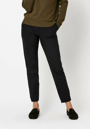 Schwarze maßgeschneiderte Hosen zeichnen sich durch einen schlanken Schnitt, eine glatte Textur und einen mittelhohen Bund aus. Kombiniert mit grünen Loafers und einem olivgrünen Pullover.
