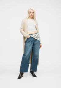 Γυναίκα με μακριά ξανθά μαλλιά φοράει μπεζ μακρύ ζακέτα, λευκό κοντό τοπ, μπλε τζιν παντελόνι φαρδιάς γραμμής, μαύρες μπότες με τακούνι και πλεκτή τσάντα μέσης.