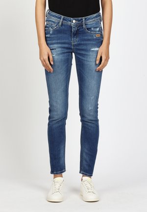 Frau trägt eng anliegende blaue Jeans mit Used-Look und weiße Sneaker, steht vor einem schlichten Hintergrund mit locker an den Seiten hängenden Händen.