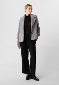 Giacca esterna di un leggero viola con texture, colletto a risvolto, top nero aderente, pantaloni neri larghi e stivali neri a metà polpaccio.