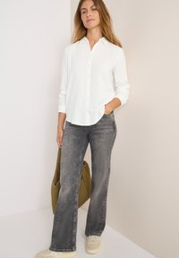 Chemise blanche à boutons avec des manches longues, associée à un jean évasé gris. La tenue comprend un sac vert et des baskets beiges. L'arrière-plan est neutre.