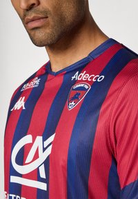 Raudonai mėlynos juostelės futbolo marškiniai su V formos kaklu. Yra logotipai: "Kappa", "Adecco" ir "Clermont Foot". Audinys tekstūruotas, puikiai tinkantis sportui.