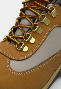 Brown leather shoe with a textured gray fabric panel, yellow and maroon laces, and gold metal eyelets. White stitching accents the design.
