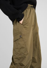 Olijfgroene cargobroek met een elastische tailleband, zijzakken en een knoopdetail op de zak. Gemaakt van lichte, gestructureerde stof.