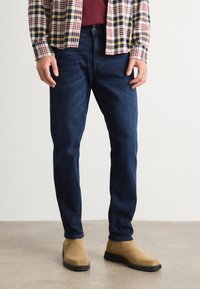 Blue denim jeans with a regular fit, paired with a plaid shirt in red, yellow, and navy, accessorized with tan suede boots.