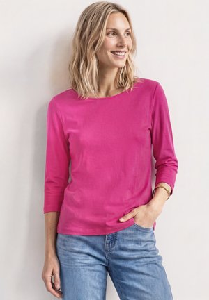 Femme aux cheveux blonds mi-longs, portant un t-shirt rose vif à manches longues et un jean bleu clair, souriante et regardant sur le côté.