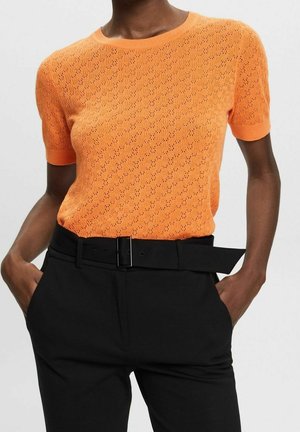 Orange gestrickter Pullover mit kurzen Ärmeln und strukturiertem Muster, kombiniert mit schwarzen Hosen, die einen Gürtel und seitliche Taschen haben.