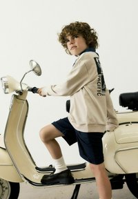 Sweatshirt beige clair avec détail graphique, short marine, chaussettes blanches et chaussures noires ; assis sur une trottinette crème avec des accents chromés.