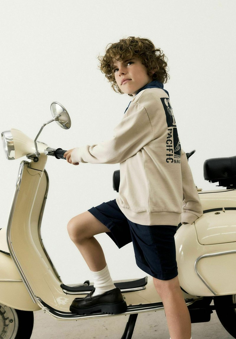 Sweatshirt beige clair avec détail graphique, short marine, chaussettes blanches et chaussures noires ; assis sur une trottinette crème avec des accents chromés.