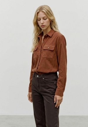Jeune femme aux longs cheveux blonds portant une chemise couleur rouille avec boutons et un pantalon marron foncé, debout devant un fond clair uni.