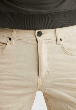 Close-up van beige spijkerbroek met een zwarte knoop en klinknagels, gedragen met een donker geribd topje met tailleband.