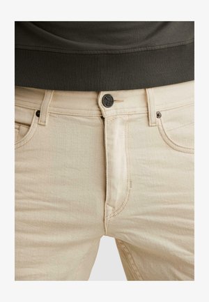 Close-up van beige spijkerbroek met een zwarte knoop en klinknagels, gedragen met een donker geribd topje met tailleband.