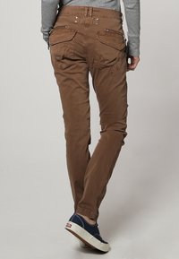 Brown cotton trousers with a slim fit, featuring two back pockets and zipper details; paired with navy canvas shoes.