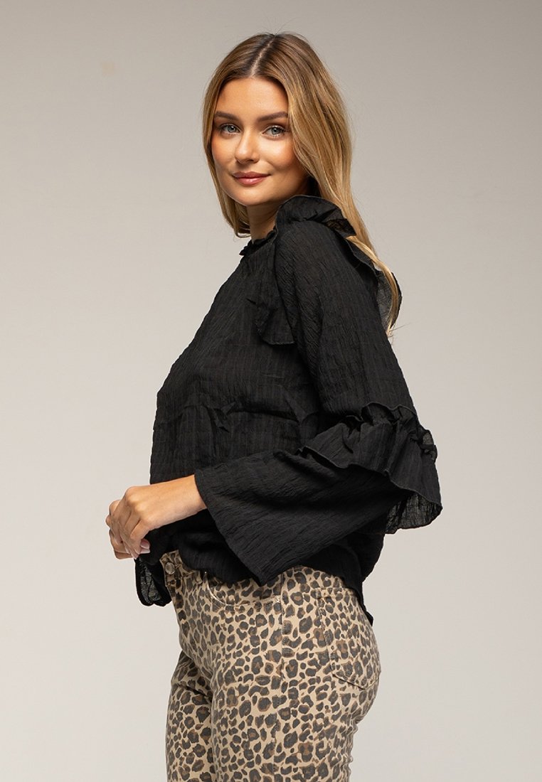 Schwarze Bluse mit strukturiertem Stoff und Rüschendetails an den Ärmeln, kombiniert mit hellbraunen Tierdruckhosen im Slim Fit.